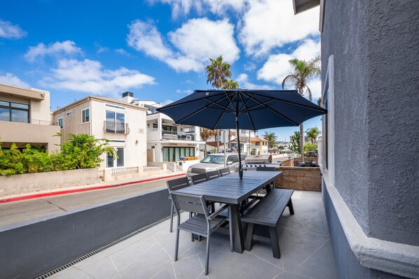 An outdoor patio with a table and chairs, a large umbrella, and a view of the street with buildings and palm trees.