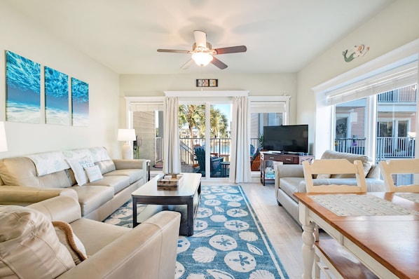 A living room with a sofa, coffee table, and TV. There is a balcony with a view of palm trees and a building. - Gorgeous 3 bedroom, 3 bath townhome with stunning water views! (Pensacola Beach)