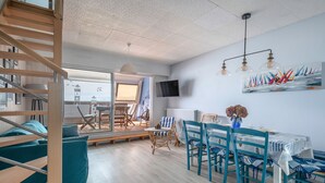 Een modern interieur met een houten trap, een eetgedeelte met blauwe stoelen en uitzicht op zee door grote ramen.