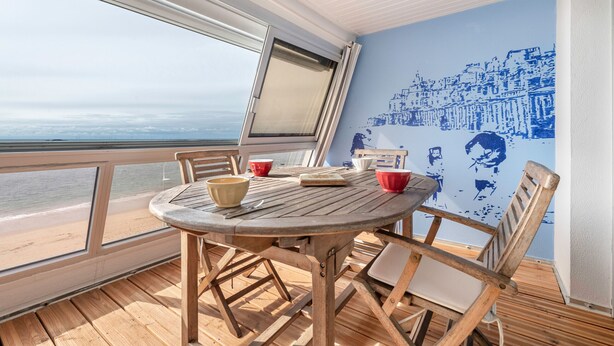 Een houten terras met een ronde tafel en stoelen, met uitzicht op een strand.
