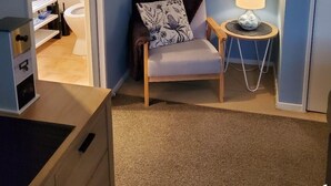 A room with a blue wall, a framed picture, a chair with a floral pillow, a small table with a lamp, and a door leading to another room. - Suburbia East Auckland (Auckland)