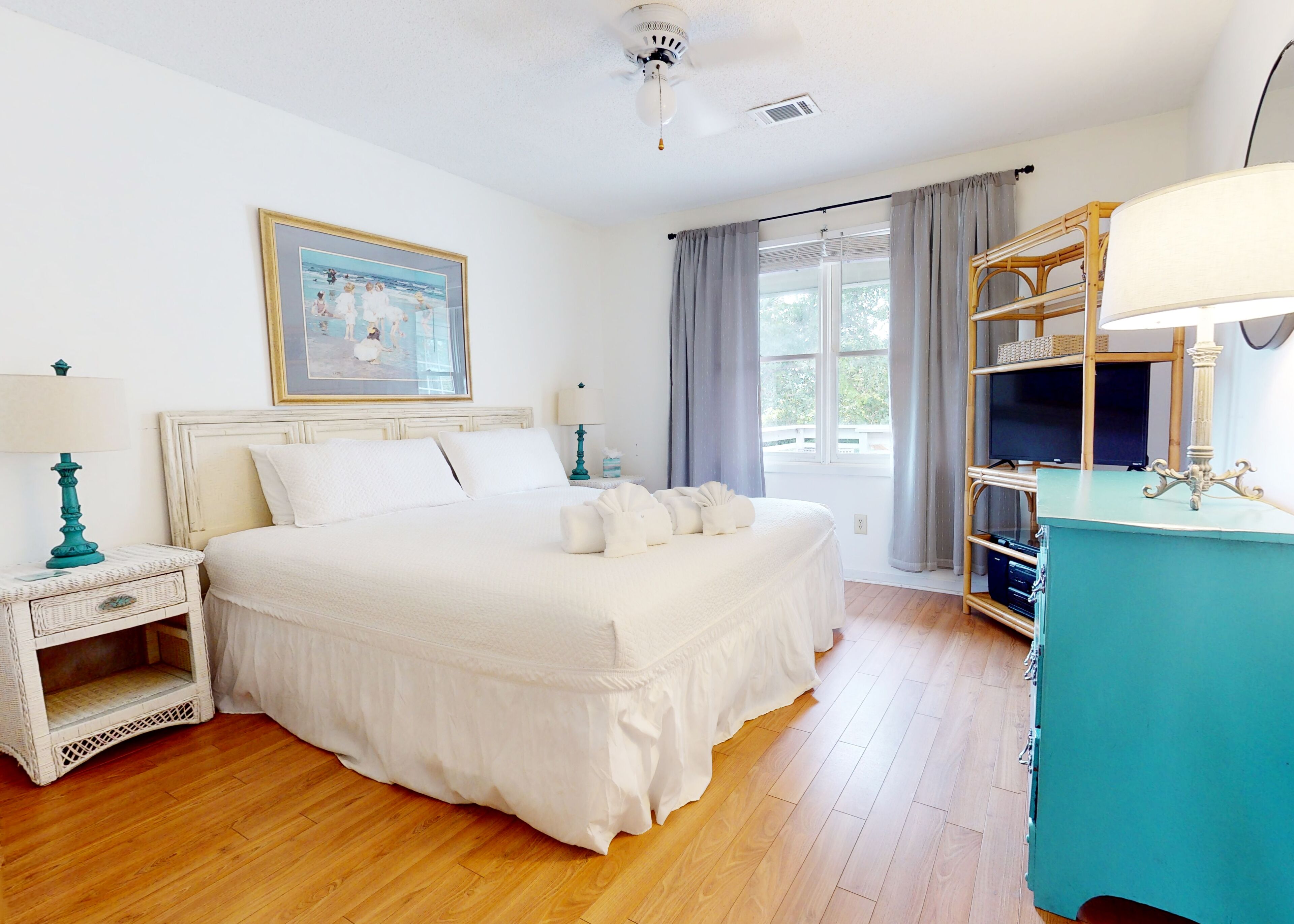 A bedroom with a large bed, a ceiling fan, a window with curtains, a television, and a small table with a lamp.