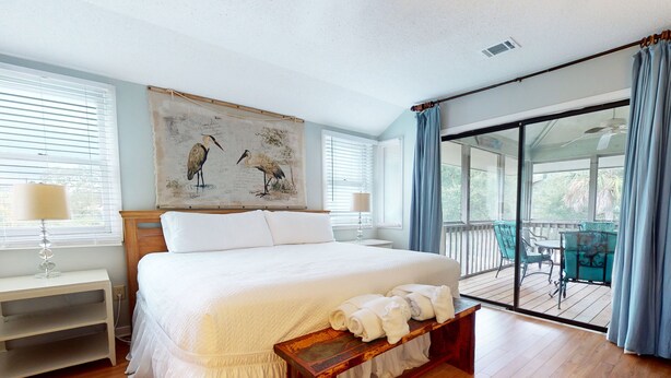 Un dormitorio amplio con una cama grande, suelo de madera, un balcón con cortinas azules y un cuadro de pájaros en la pared.