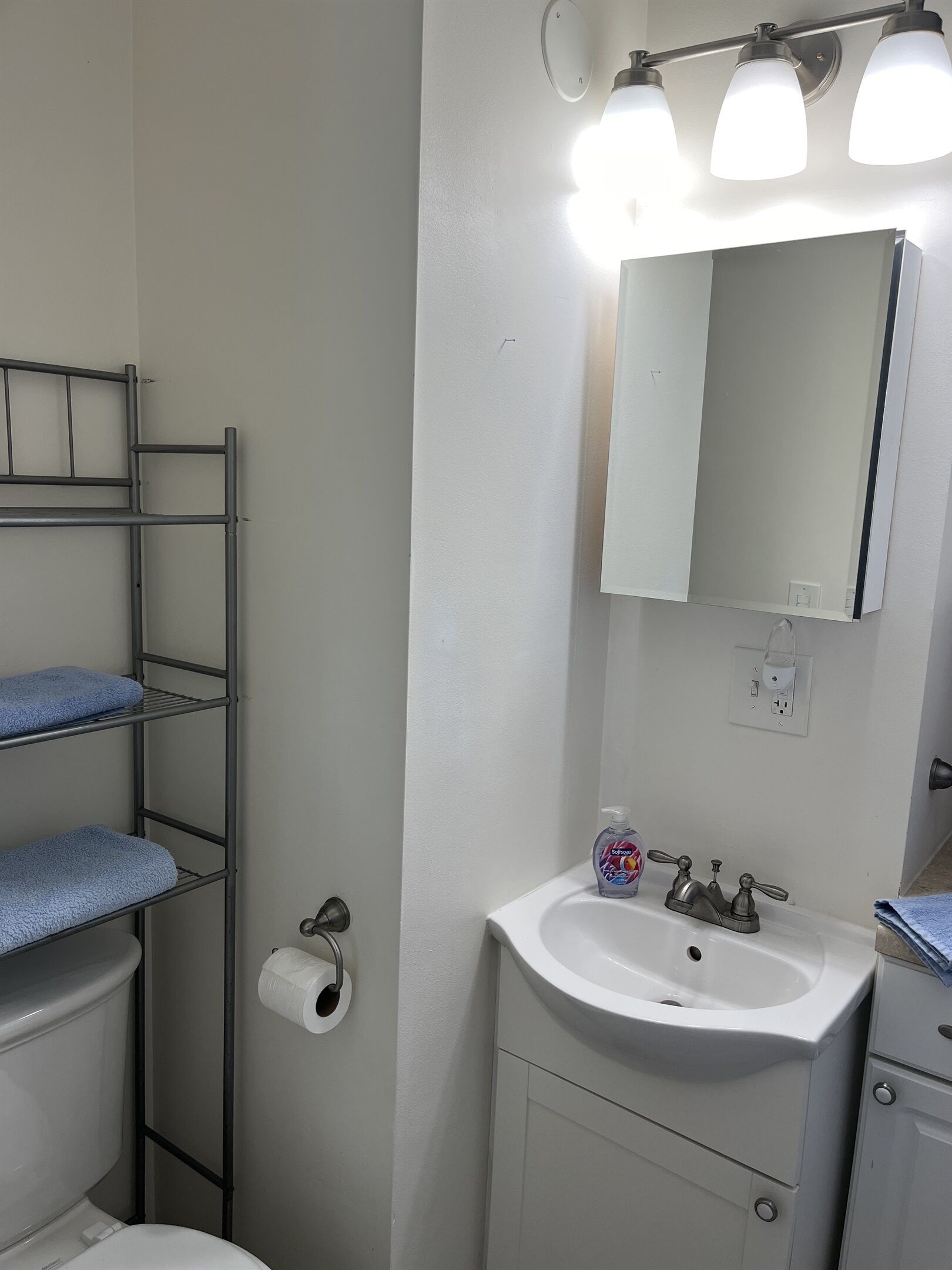 Een badkamer met een wit wastafel, een spiegel en een toilet. Er is een metalen plank met handdoeken en een toiletrol.