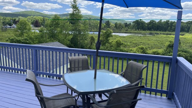 Um terraço com mesa e cadeiras de vidro, um guarda-sol azul e vista para um lago e colinas.