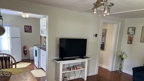 Uma sala de estar com televisão em móvel, uma mesa de jantar com jogos americanos, uma cozinha com eletrodomésticos e um corredor que dá para outro quarto.