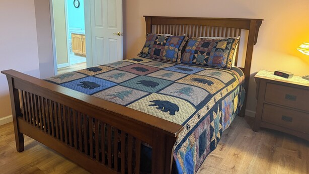A bed with a quilt, a wooden headboard, a nightstand with a lamp, and a door with a clock. - Grandview Private 2 room suite, 5 minutes to Dreams Park, close to town (Milford)