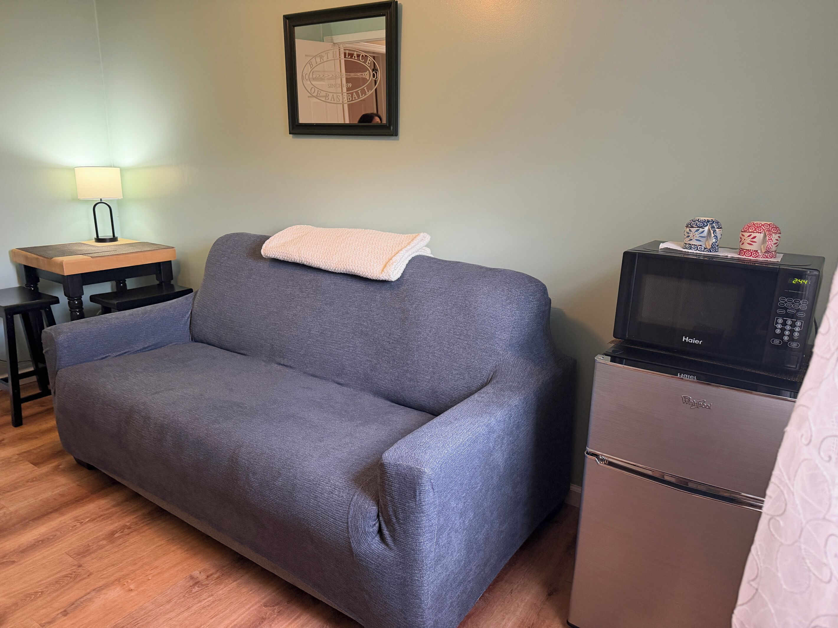 A room with a grey sofa, a small table with a lamp, a microwave, and a framed picture on the wall.
