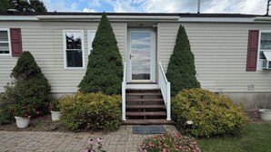 A single-story mobile home with a front porch, two potted evergreen trees, and a flower bed with pink flowers. - Grandview Private 2 room suite, 5 minutes to Dreams Park, close to town (Milford)