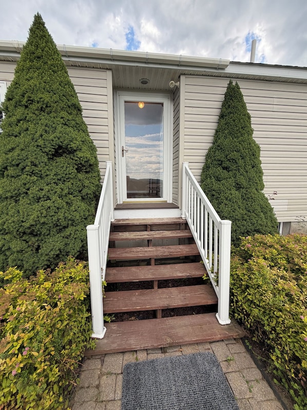 A set of wooden stairs leading to a white door, flanked by two coniferous trees and a small garden area with a doormat.