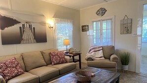Een woonkamer met een beige bank, een salontafel en een stoel. Er staat een lamp op een bijzettafeltje, een schilderij hangt aan de muur, en er is een raam met zonwering.