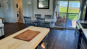 A modern kitchen with a central island, a dining area with chairs, a built-in washer and dryer, and a view of a green landscape through a sliding glass door. - Grousehall Total Renovation, contemporary design, short drive to Cooperstown (Schuyler Lake)
