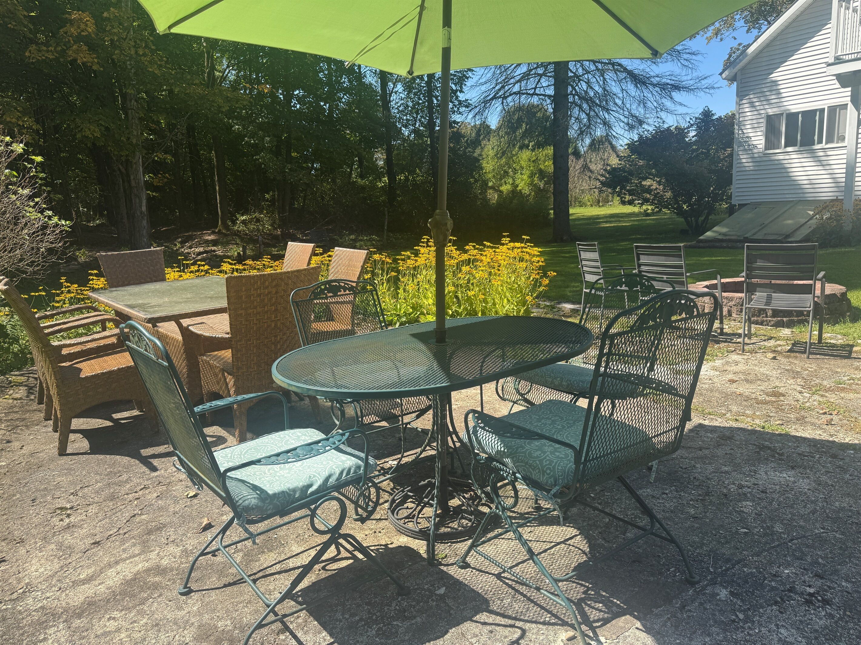 Een terras buiten met een ronde tafel en stoelen, omringd door rieten meubels en een groene parasol.