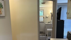 Una cocina con un lavamanos de acero inoxidable, una ventana con persianas y una puerta que da a un baño con inodoro y lavamanos.