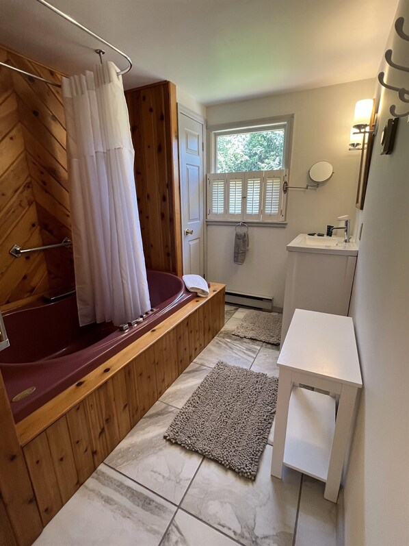 A bathroom with a wooden vanity, a bathtub, a window with shutters, and a small white table.