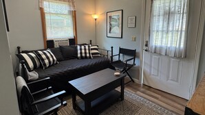 A cozy living room with a dark-colored sofa, two black chairs, a small wooden table, and a patterned rug. There is a window with blinds, a wall-mounted lamp, and a framed picture.