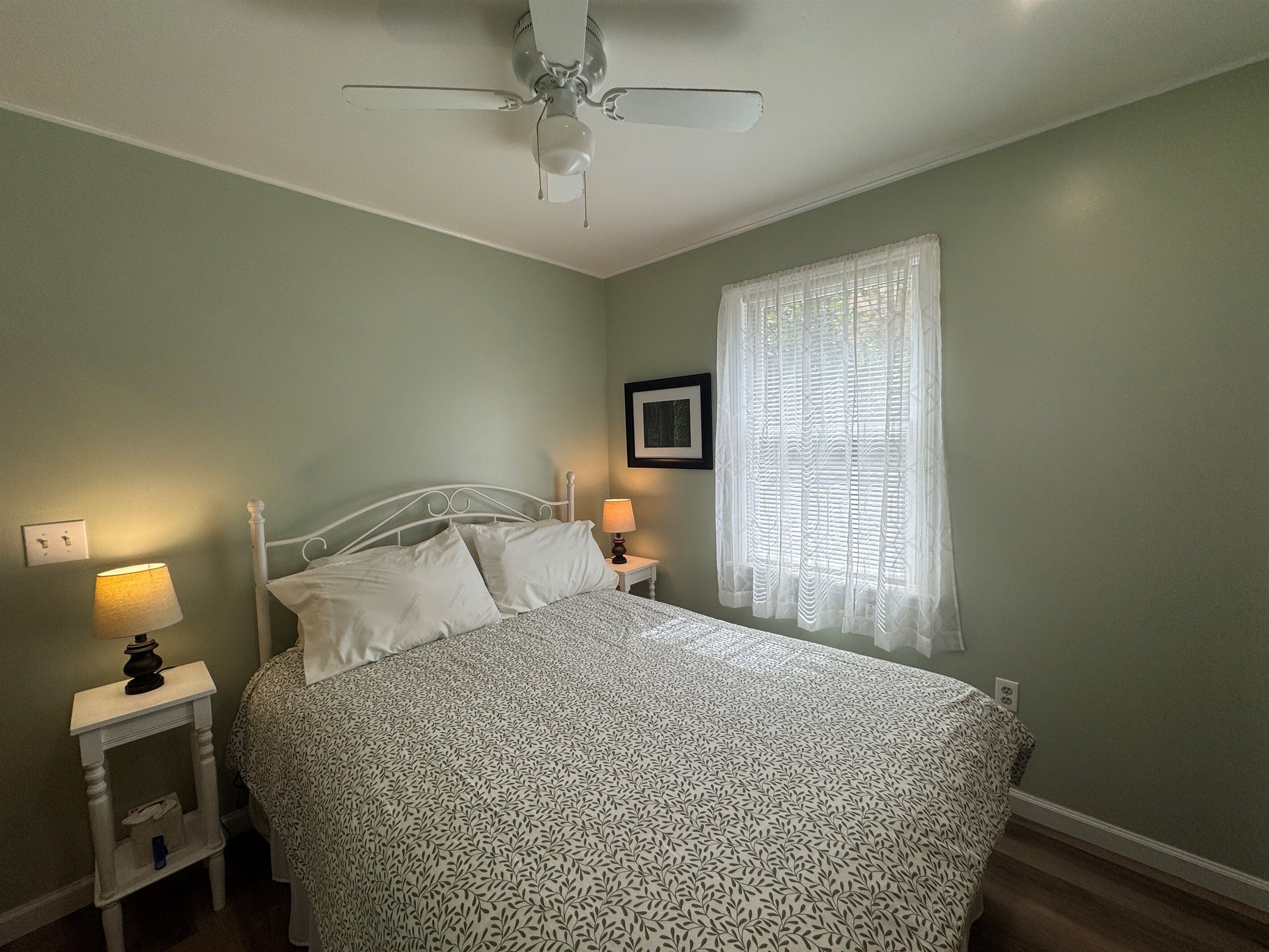 Une chambre à coucher avec un lit, deux tables de chevet avec des lampes, un ventilateur de plafond, une fenêtre avec des rideaux légers et un tableau encadré au mur.