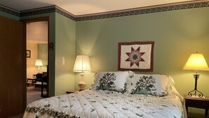 A bedroom with a floral bedspread, two bedside lamps, a decorative wall hanging, and a door leading to another room. - Morgan Farm - Well cared for Ranch house near Canadarago Lake (Richfield Springs)