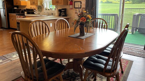 A dining area with a wooden table and chairs, a vase with flowers, and a kitchen in the background. - Morgan Farm - Well cared for Ranch house near Canadarago Lake (Richfield Springs)
