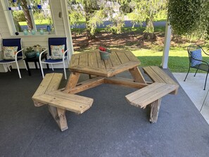 Una mesa de picnic de madera con bancos, rodeada de sillas con cojines y vistas a un jardín con árboles y césped.