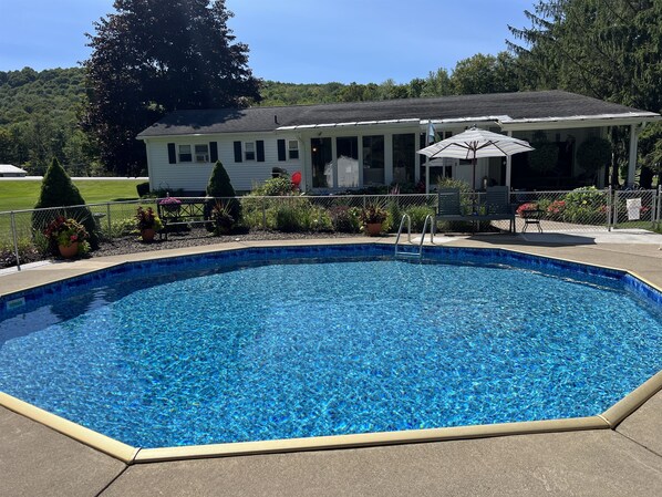 Una piscina rectangular con borde de azulejos, rodeada por una solana de hormigón. Hay un porche cubierto con una mesa y sillas, y una sombrilla.