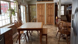 Une cuisine avec une grande table en bois, deux chaises en bois, un réfrigérateur et une fenêtre agrémentée d’une plante suspendue.