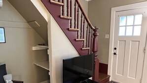 A living room with a wooden staircase, a television on a black stand, a grey couch, and a patterned rug. - Pioneer Cottage Pristine 1 bedroom home, 5 minutes to All Star, private patio (Oneonta)