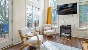 A living room with two light-colored armchairs, a wooden coffee table, a flat-screen TV mounted on the wall, and a large window with a view of trees. - Updated Flooring, New Hot Tub, Resort Amenities! (Mc Gaheysville)
