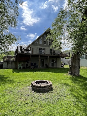 A two-story house with a balcony, a fire pit in the front yard, and a large tree to the right.
