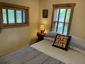 A bedroom with a bed, two windows, a nightstand with a lamp, and a framed picture on the wall.