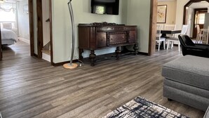 Uma sala de estar com piso de madeira, um armário de madeira escura, um abajur moderno, uma TV de tela plana fixada na parede e um sofá cinza.