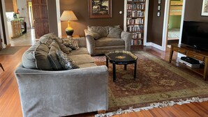 A living room with a grey sofa, a brown armchair, a coffee table, a bookshelf, and a television.