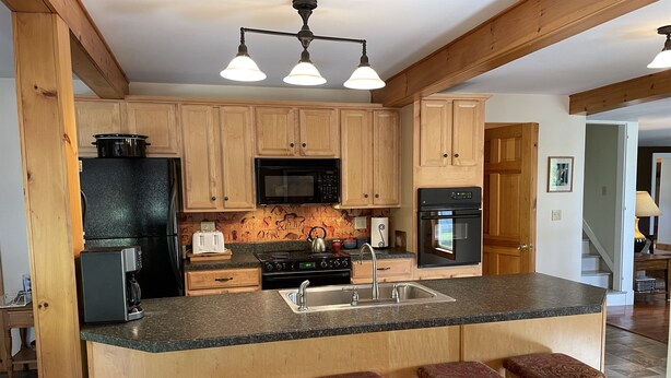 A kitchen with wooden cabinets, a black refrigerator, a microwave, a stove, and a sink. There are three stools with patterned cushions.