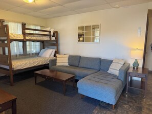 A bunk bed with a patterned bedspread, a grey sofa with striped cushions, a small wooden table, and a nightstand with a lamp. - Sunnyside Apartment 1#, 1 of 3 - Great location, pool, 1/2 mile to Dreams Park (Cooperstown)