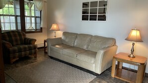 Een woonkamer met een beige bank, een geruit fauteuil, twee lampen, een raam met gordijnen en een klein tafeltje.
