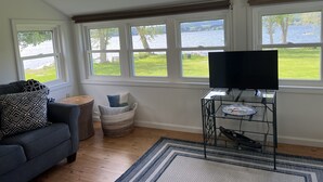 Een woonkamer met een bank, een televisie op een meubel, een raam met uitzicht op een meer, en een klein tafeltje met een mand.