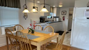 Een compacte keuken met een eethoek, houten stoelen, een centrale tafel met een bloevas, hanglampen en een witte koelkast.