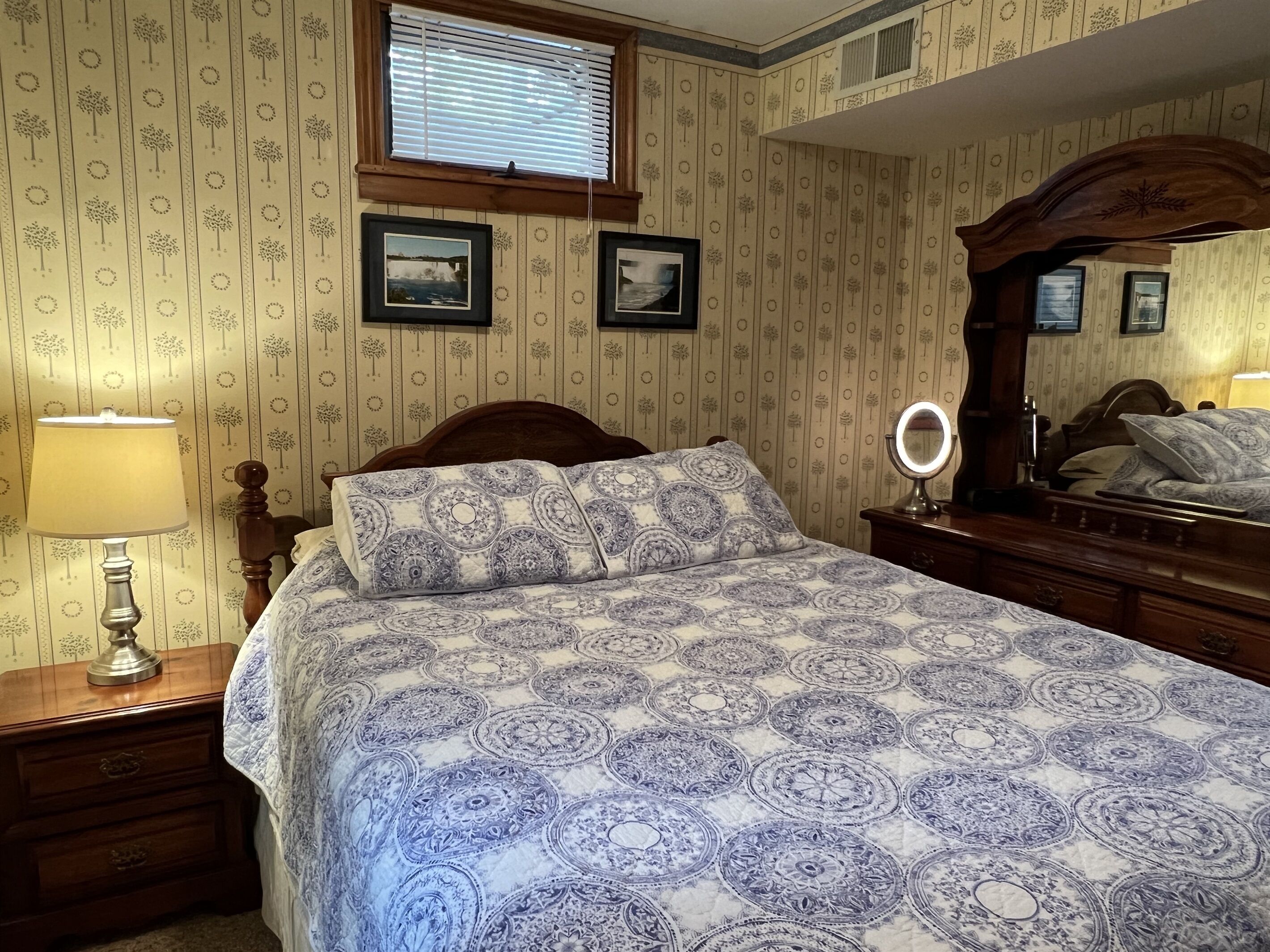 Un dormitorio con una cama, una mesita de noche, un tocador, un espejo y dos cuadros en la pared.