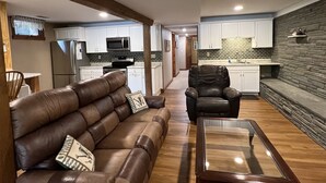 A modern living room with a leather sofa, a glass coffee table, and a leather armchair. A kitchen with white cabinets and a stone accent wall is visible in the background. - Sunset Waters -Goodyear Lake - hot tub, swimming ,fishing. paddle sports (Maryland)