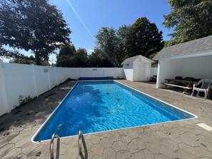 Piscina rectangular con una cerca blanca alrededor, un cobertizo pequeño y una mesa de picnic con sillas.