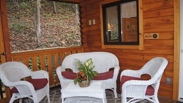 Eine Holzdiele mit Veranda, ausgestattet mit Korbstühlen und einem Tisch, ein Fenster mit Blick auf einen Wald und eine Wandplakette.