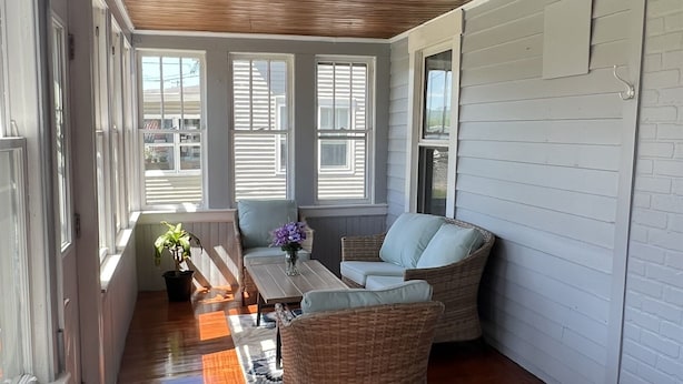 Une pièce lumineuse, avec un plancher en bois, un ventilateur de plafond et un coin salon comprenant des fauteuils en osier et une table.