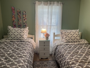 Un dormitorio con dos camas, una mesita de noche con lámpara y una ventana con cortinas traslúcidas.