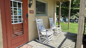 A covered porch with a red door, two white rocking chairs, a stone floor, and a view of a backyard with outdoor furniture.