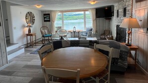 Una habitación acogedora con una mesa redonda de madera, dos mecedoras, una chimenea y un ventanal grande con vistas a un lago.