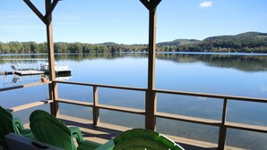 Una terraza de madera con sillas verdes que da a un lago tranquilo, barcos amarrados y una vista despejada de las colinas circundantes.