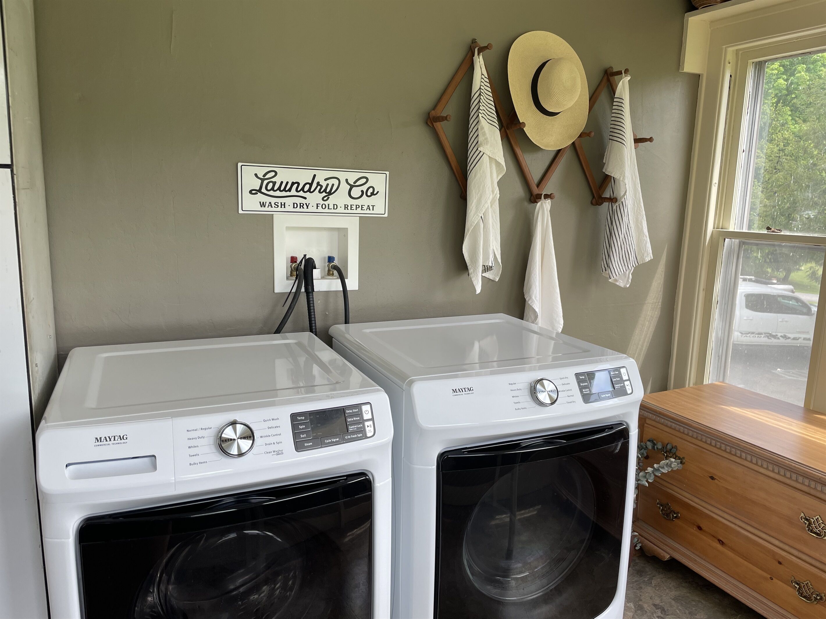 A laundry area with two Maytag washing machines, a sign reading 'Laundry Co Wash Dry Fold Repeat,' and a window with a view of a car outside.