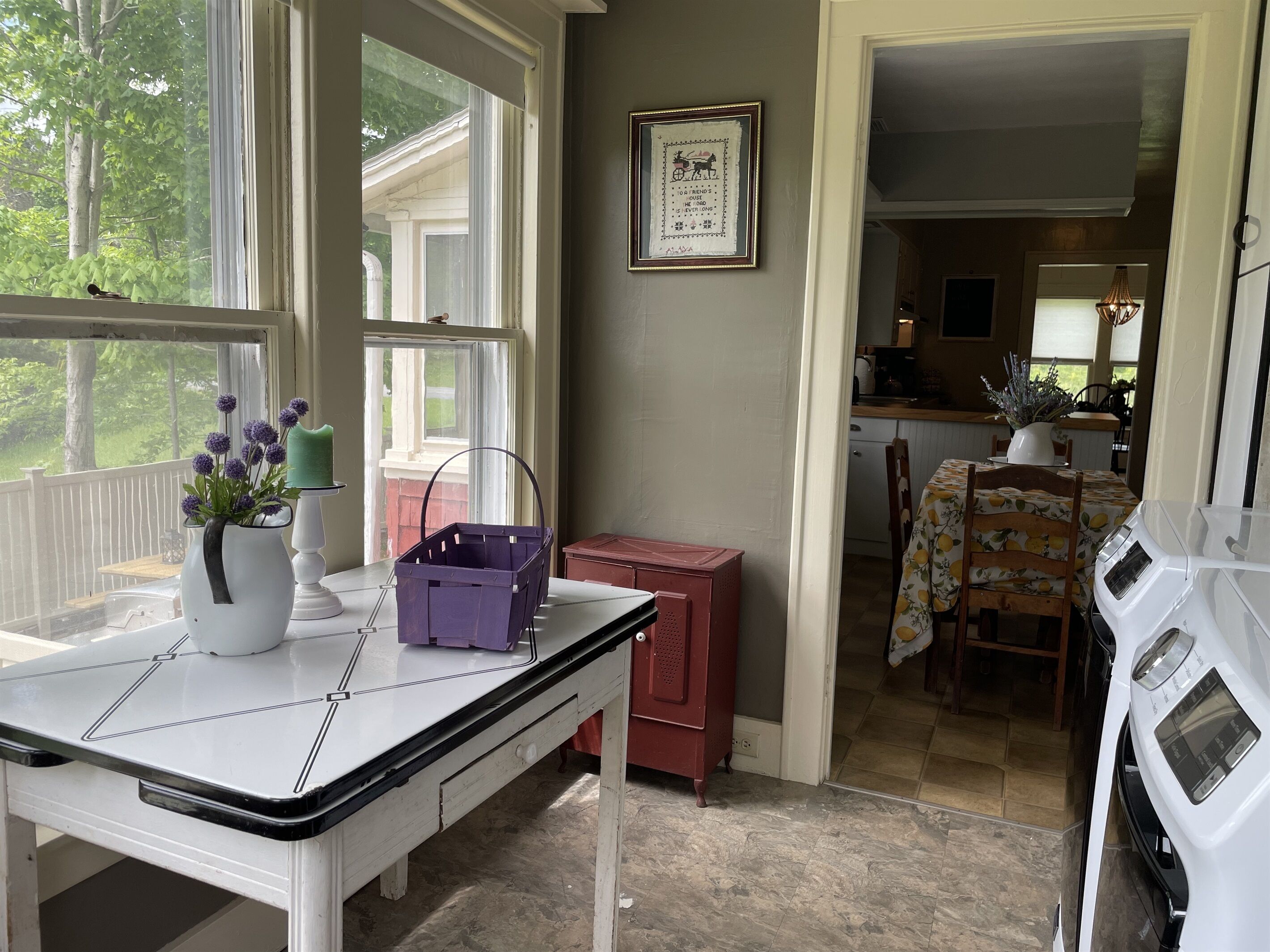 Una cocina con una mesa de comedor, una cesta morada, una jarra blanca con flores y un cuadro en la pared.