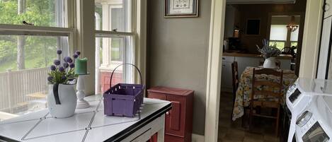 Una cocina con una mesa de comedor, una cesta morada, una jarra blanca con flores y un cuadro en la pared.