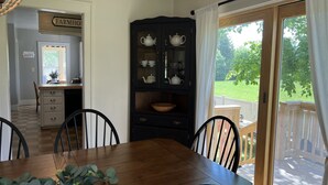 A dining area with a wooden table and chairs, a dark cabinet with glass doors, and a view of a green landscape through a large window. - Wake Robin Cottage - Recent renovation 4 bdrm/ 4 bath home close to tournaments (Oneonta)